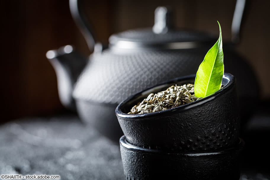 Schwarzer Teekessel im Hintergrund und schwarzer Tee in einer Schale im Vordergrund ergänzt mit grünem Schwarzteeblatt