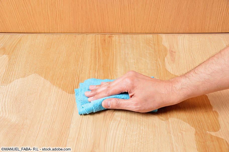 Hand mit blauem Putzlappen wischt verschüttete Flüssigkeit auf dem Parkettboden auf
