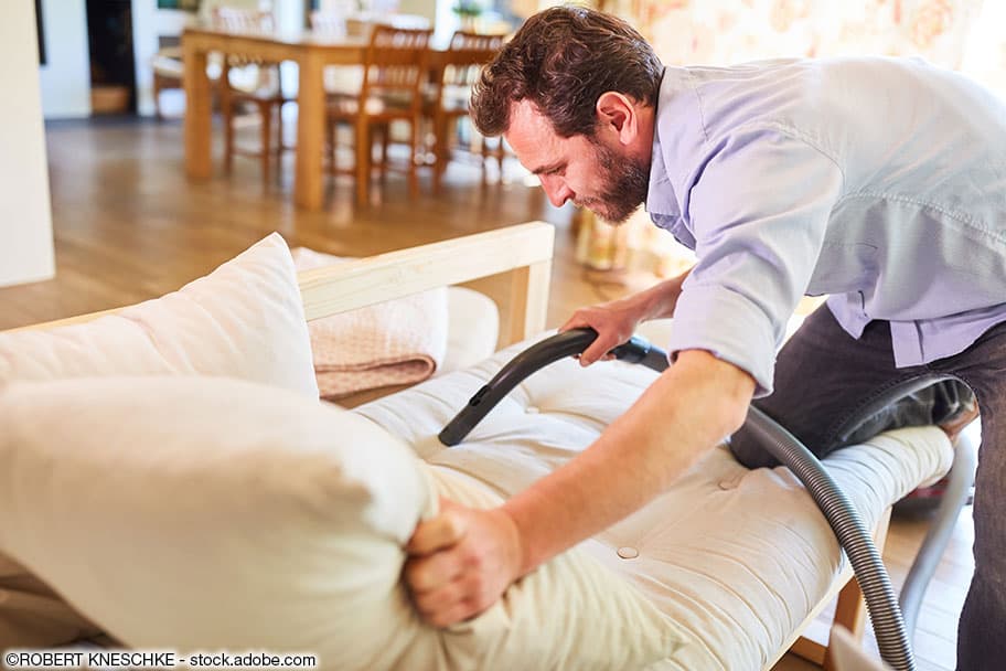 Mann saugt sein weisses Sofa mit einem Staubsauger ab. Er trägt ein violettes Hemd und graue Jeans.